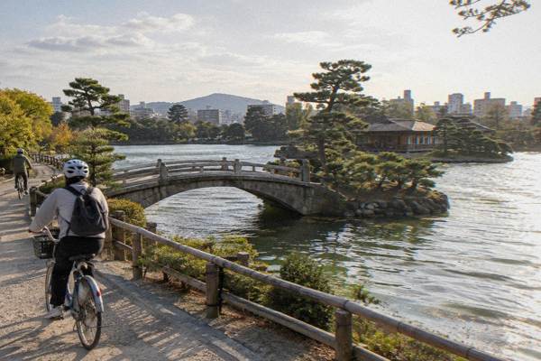 A guide leads a line of cyclists along a riverside path past trees and bridges.