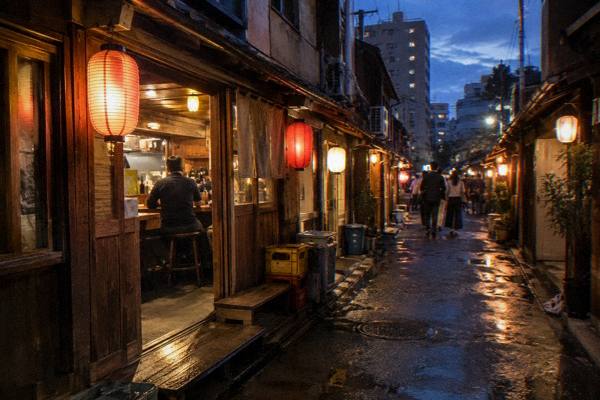 A small group follows a guide down a lantern-lit lane toward a busy izakaya counter.
