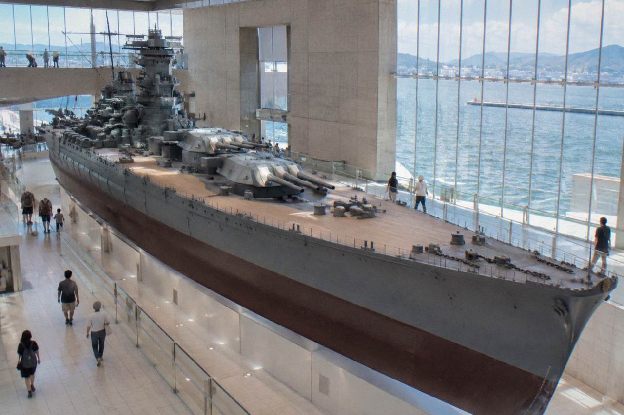 A large battleship model displayed in a bright museum atrium with visitors walking around it