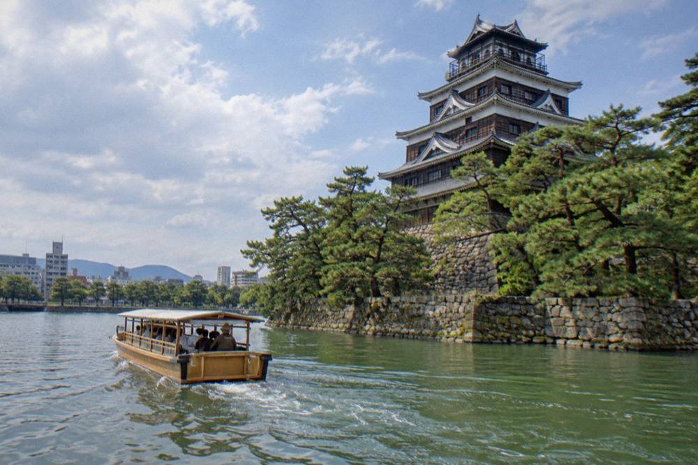 A wooden boat cruising along the inner moat of Hiroshima Castle past green pine trees