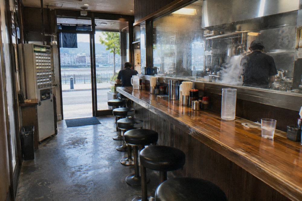 A narrow ramen counter in Hiroshima with stools, a ticket machine by the entrance and steam rising behind the glass