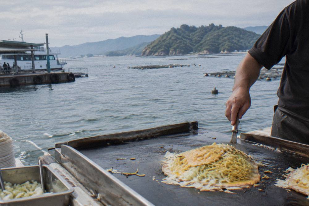 Layers of cabbage, noodles and egg stacked on a thin batter base on a griddle in Hiroshima