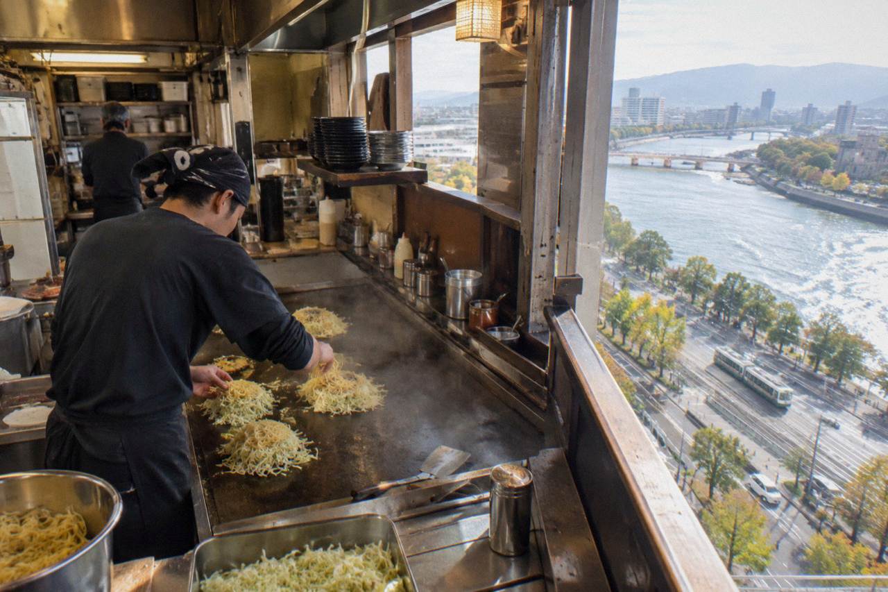 A cook layers cabbage and noodles on an iron griddle at a Hiroshima okonomiyaki counter