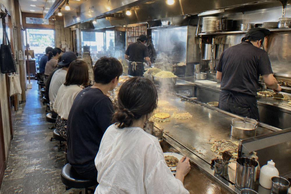 A row of customers seated at a narrow teppan counter in an okonomiyaki restaurant in Hiroshima