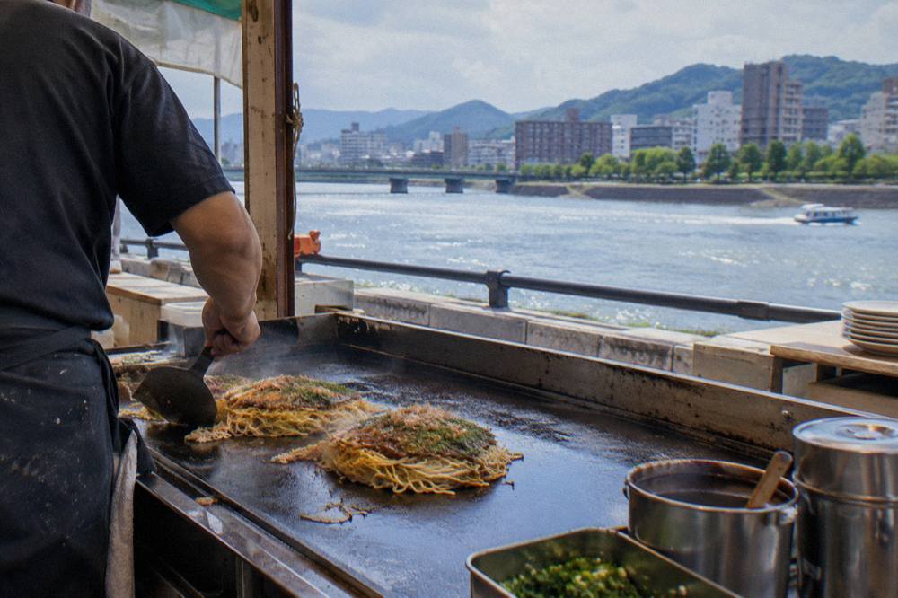 A layered Hiroshima-style okonomiyaki cooking on a flat iron griddle