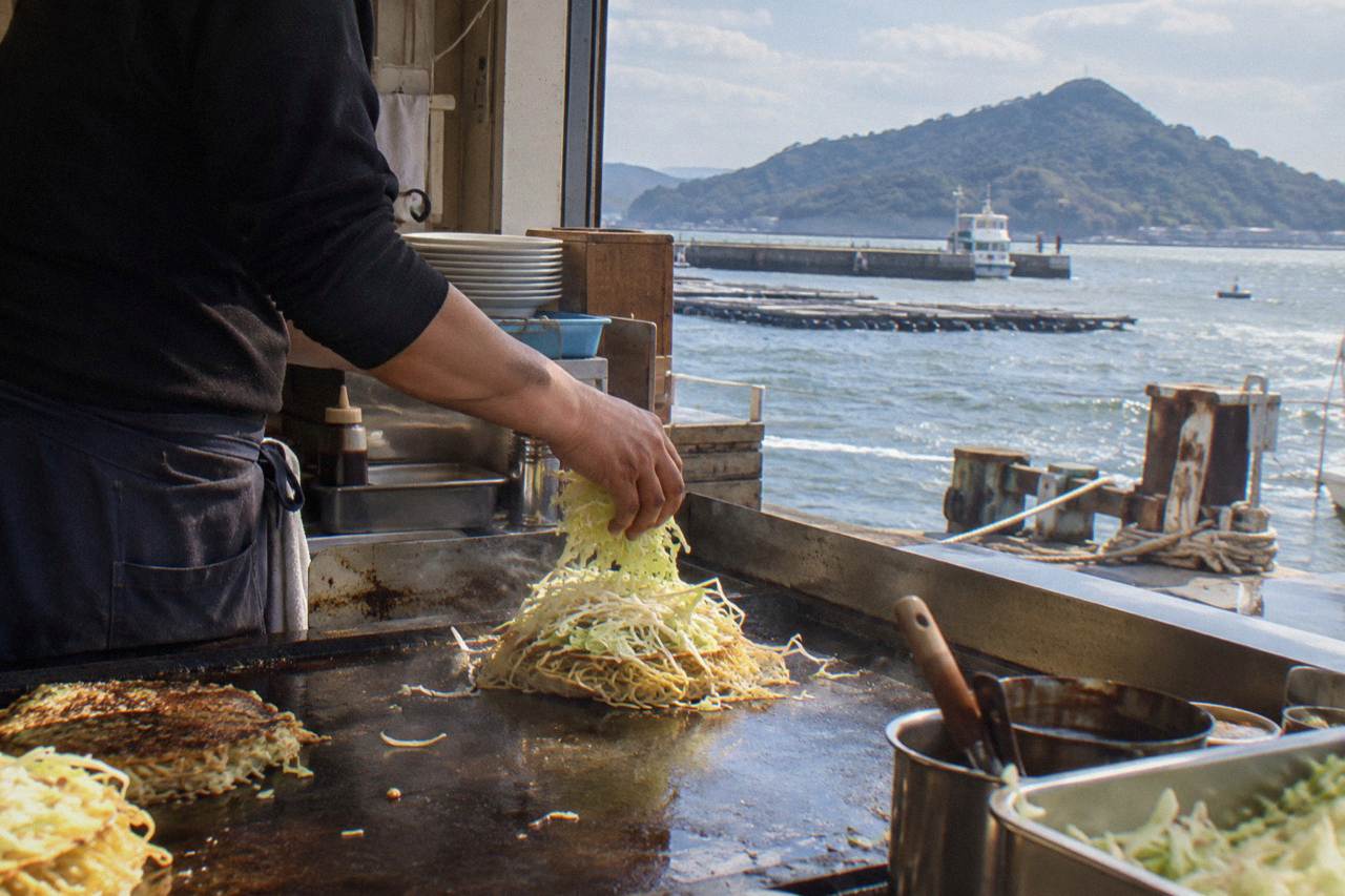 A cook layering cabbage and noodles on a sizzling okonomiyaki griddle in a small Hiroshima restaurant