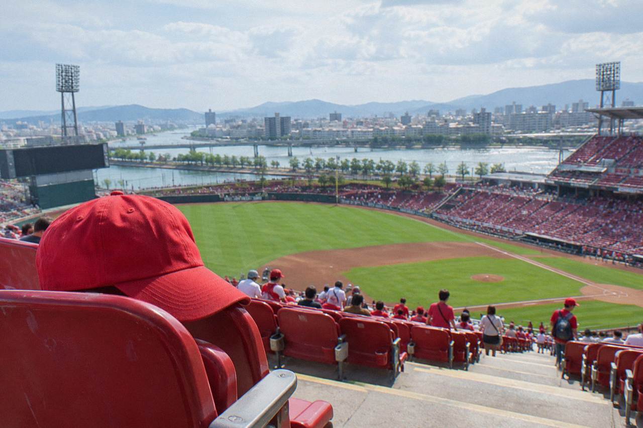 A red Hiroshima Carp baseball cap resting on a stadium seat with a ballfield in the background