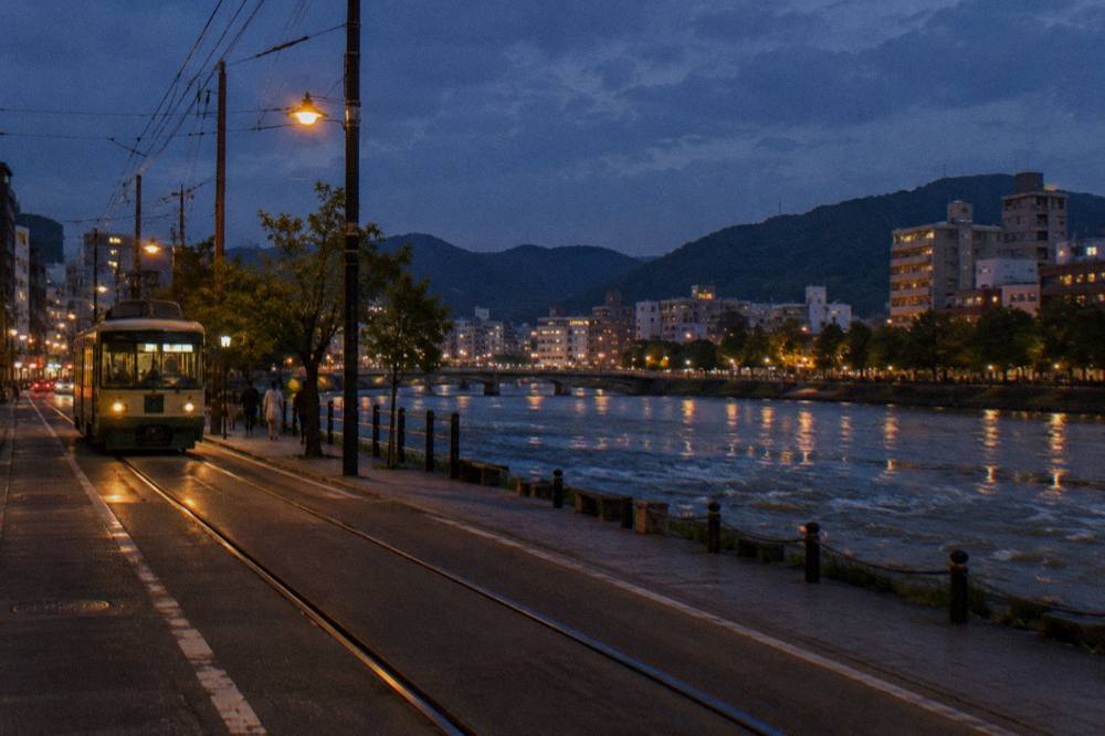 A quiet Hiroshima street scene at dusk with warm shop lights and a streetcar track running down the road