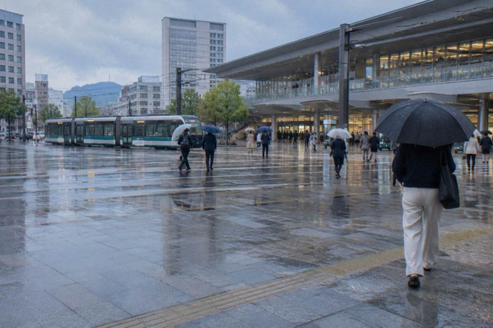 Travellers with umbrellas crossing the open plaza in front of a station entrance in rain