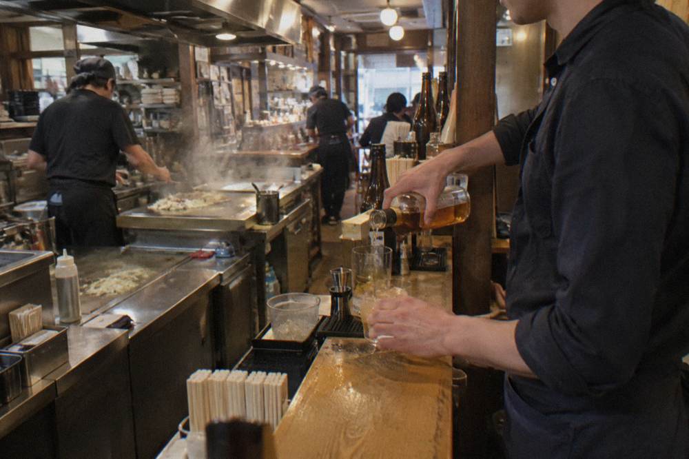 A bartender pouring Japanese whisky at a small standing bar counter in Hiroshima