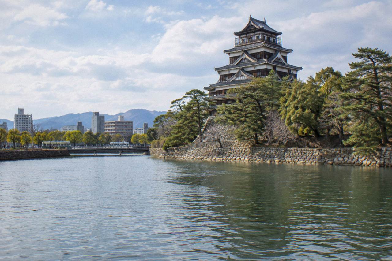 A reconstructed Japanese castle keep rising above a wide stone-walled moat, with trees along the waterline and an overcast sky.