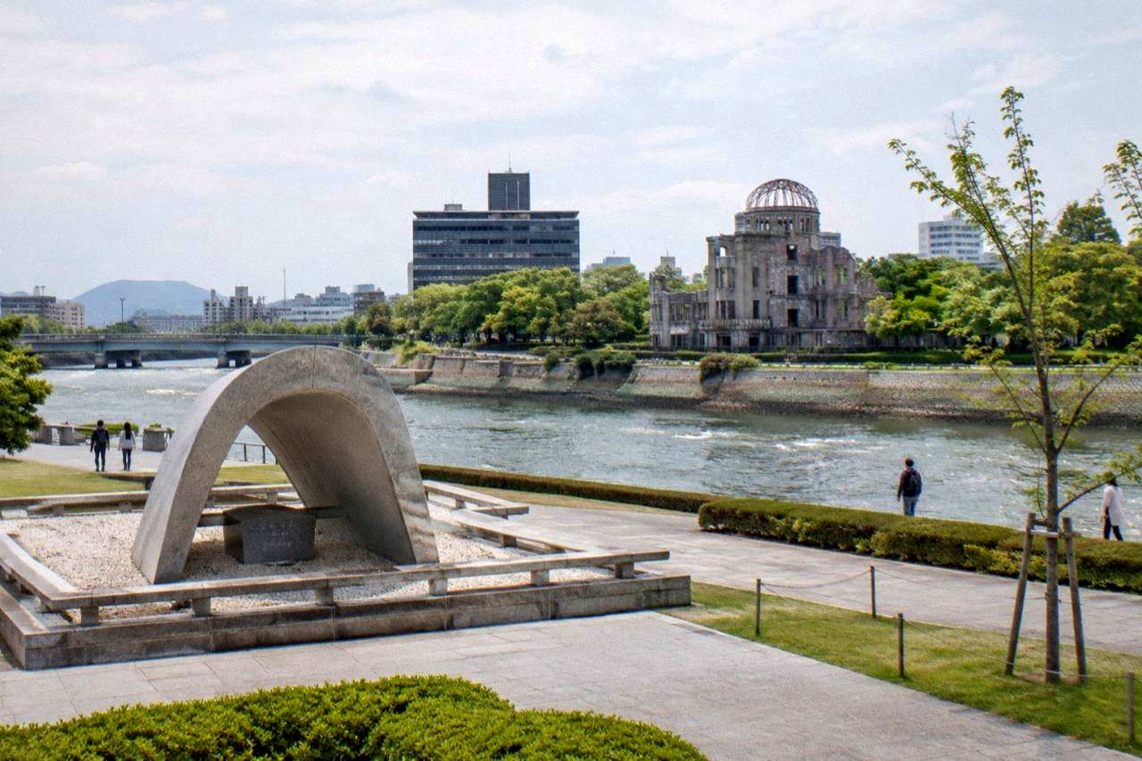 Wide view across a riverside memorial park with a flat stone cenotaph in the foreground and a domed ruin on the far bank