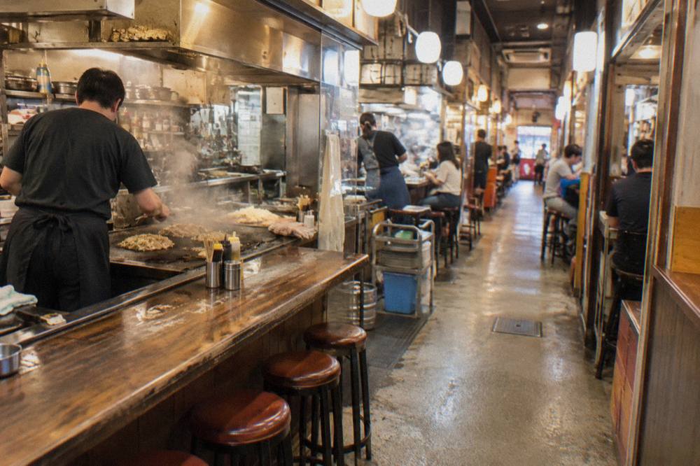 Narrow walkway inside Okonomimura food hall in Hiroshima, lined with small lit counters, steam rising as cooks work griddles