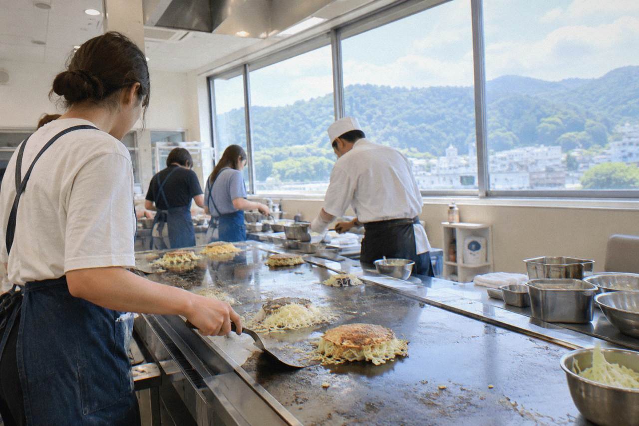 Visitors cooking okonomiyaki on a wide steel teppan griddle in a bright workshop kitchen