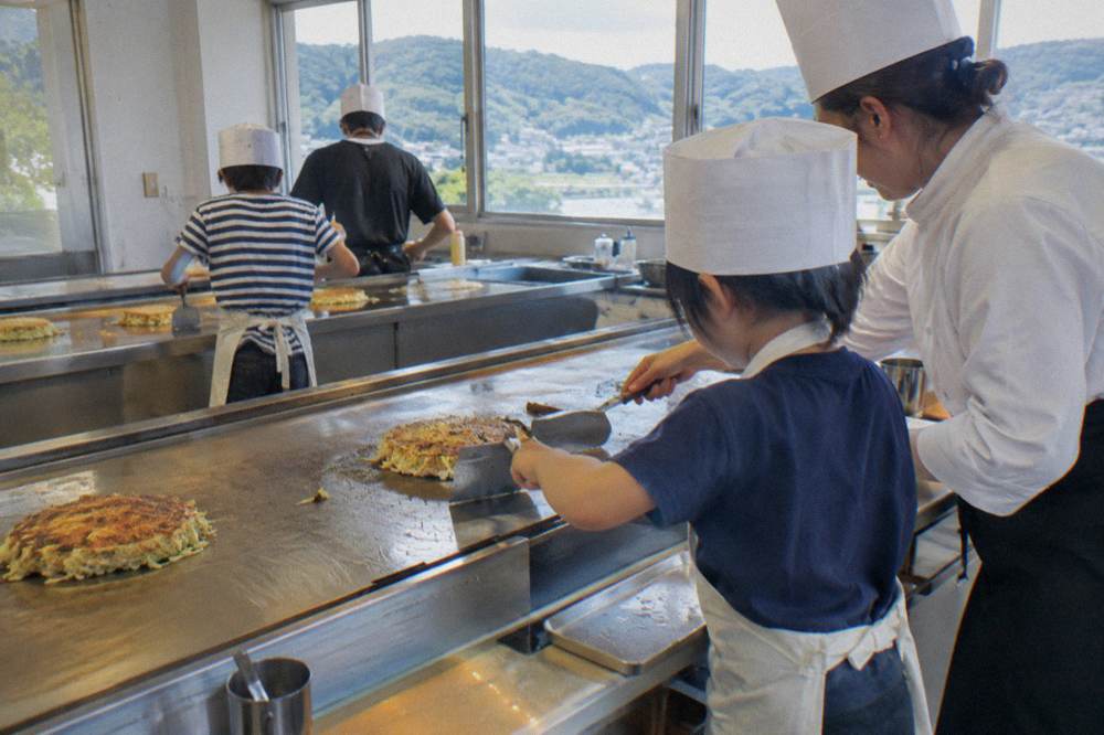 A child wearing a paper hat flipping okonomiyaki with two spatulas under a staff member's guidance