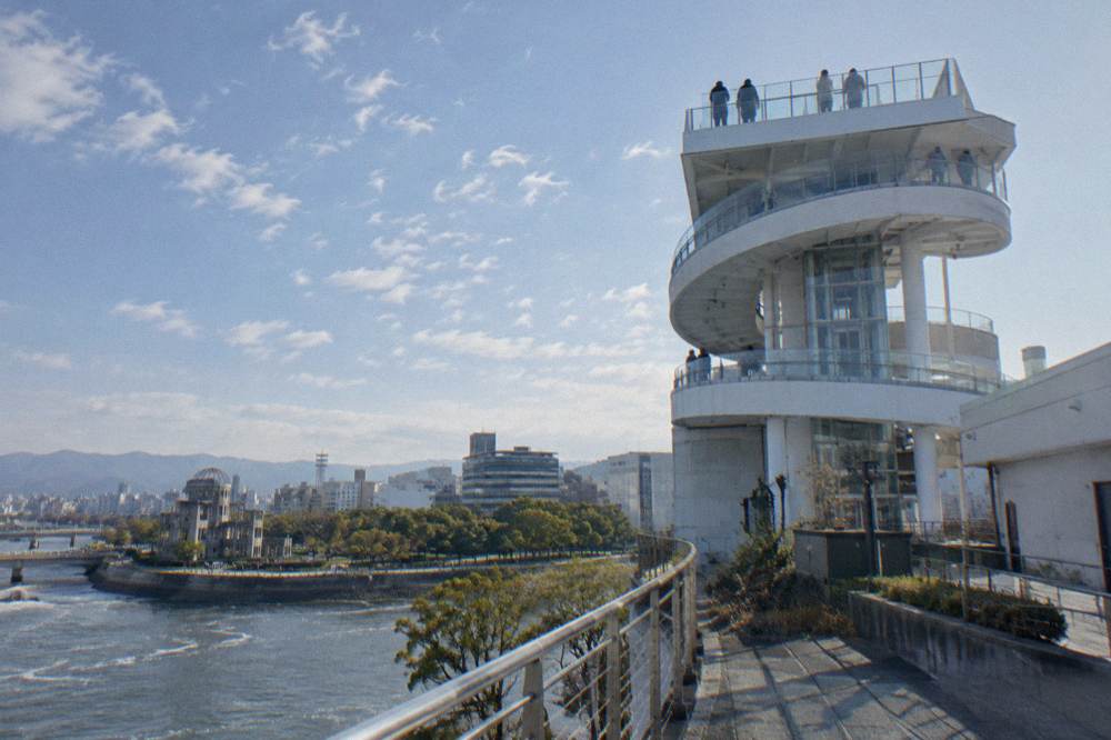 People on an open rooftop deck looking out over a river, a ruined building and city rooftops