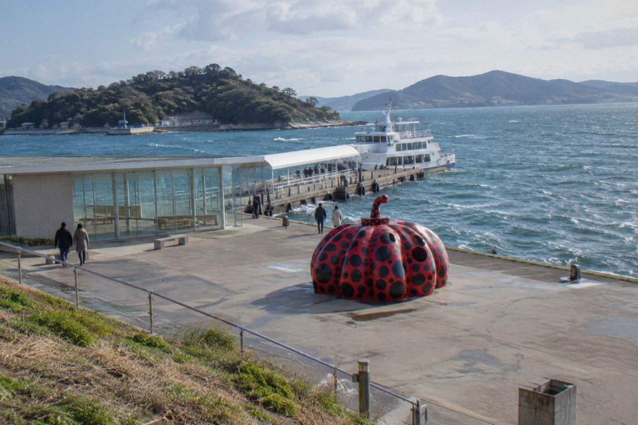 A red pumpkin sculpture on a waterfront plaza beside a ferry port on a small island