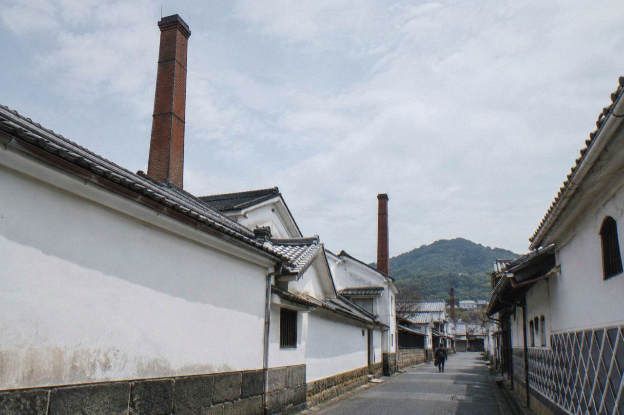 A quiet sake brewery street with white-walled brewery buildings and tall chimneys under a soft sky