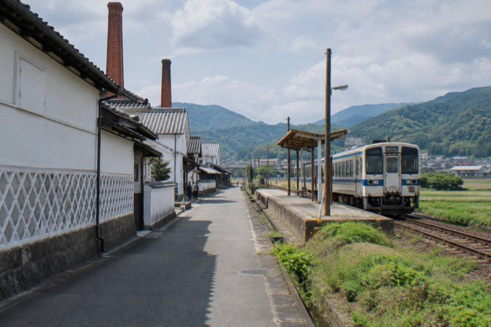 A rural train platform with a local train waiting beside fields and low hills