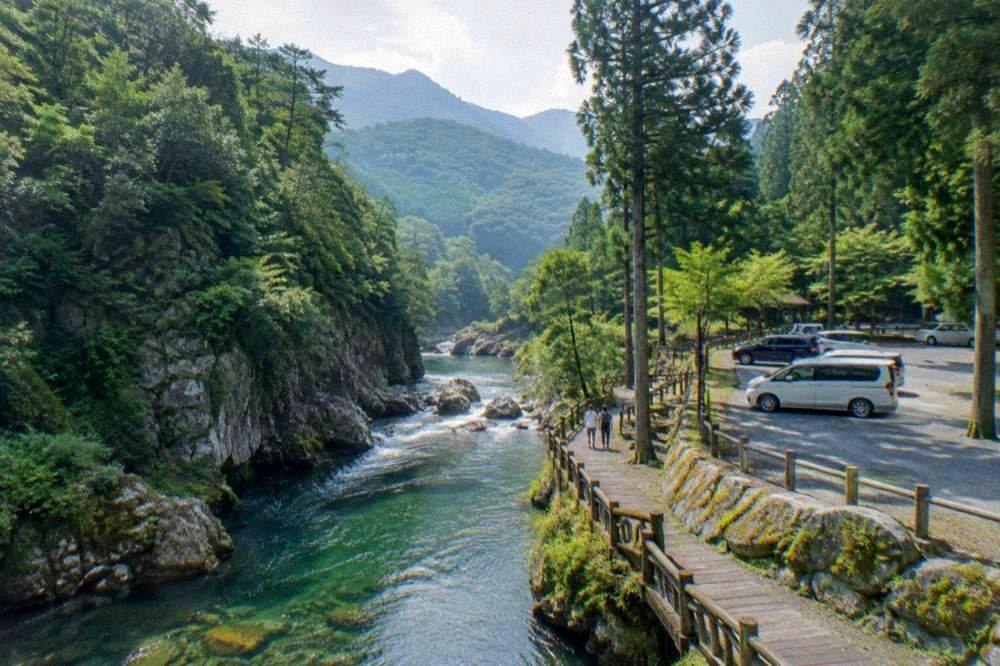 A woodland parking area beside a river with tall trees and mountains behind