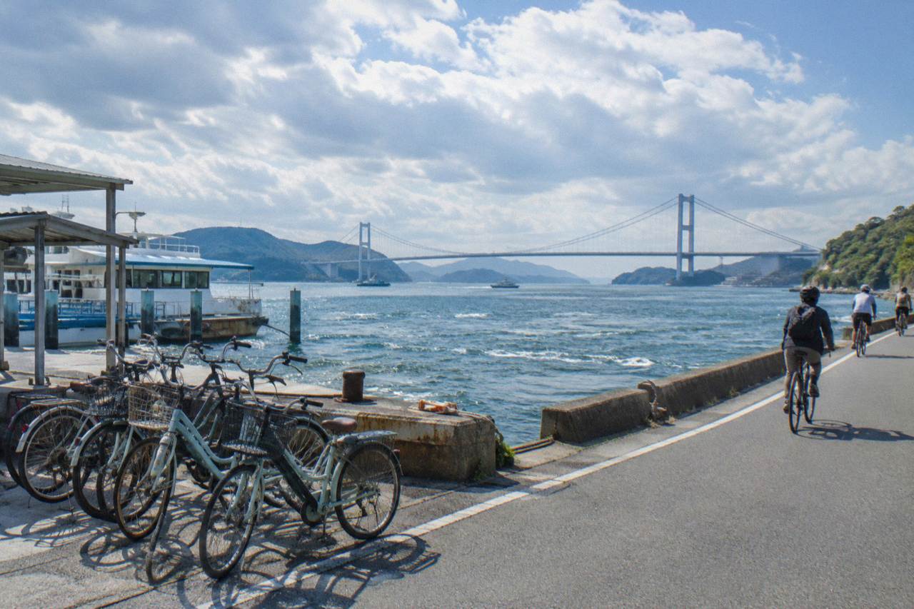 Cyclists riding along a coastal road with a long suspension bridge over blue island-dotted water
