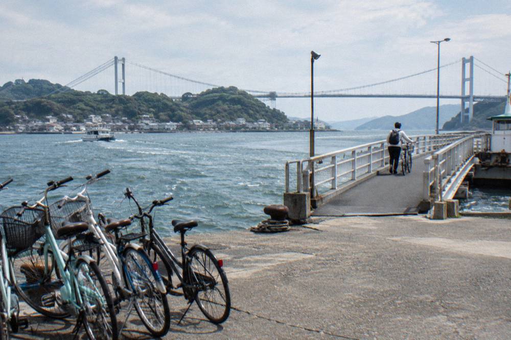 A cyclist walking a bike across a long bridge with an island town visible below