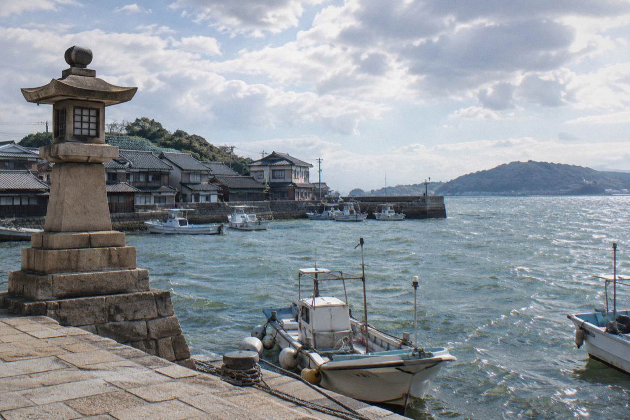 Stone lighthouse standing at the edge of a small harbor with fishing boats and old tiled rooftops behind it