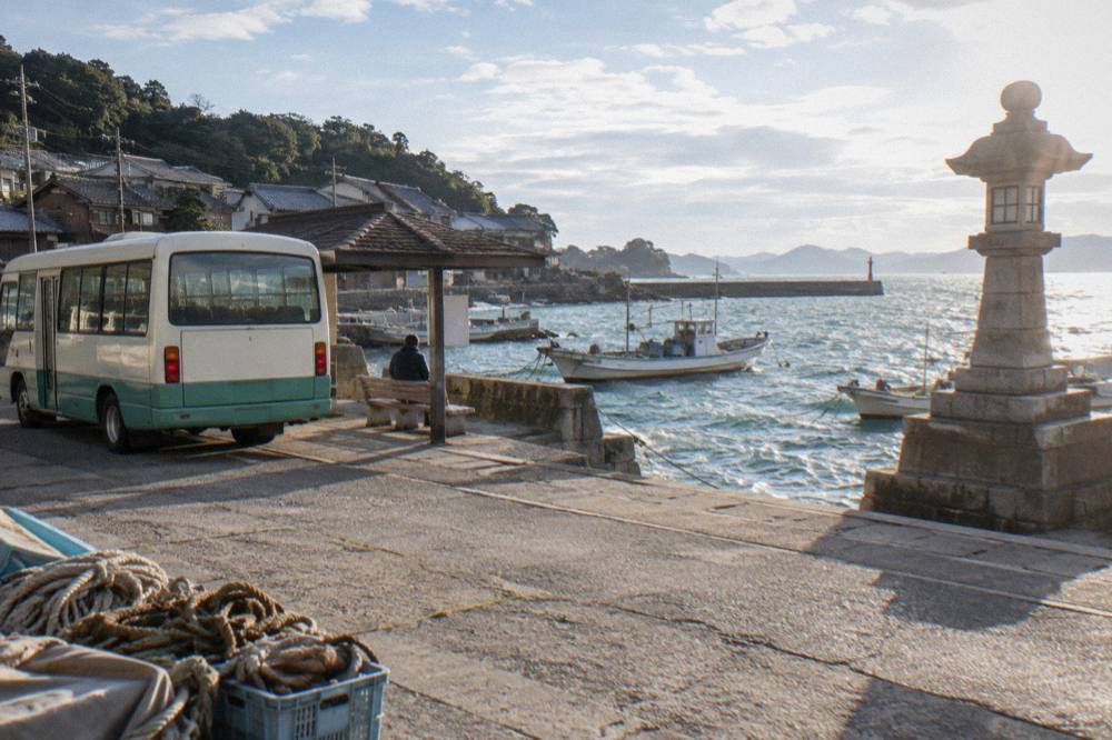 Local bus parked at a small Japanese town bus stop near a harbor with a stone wall