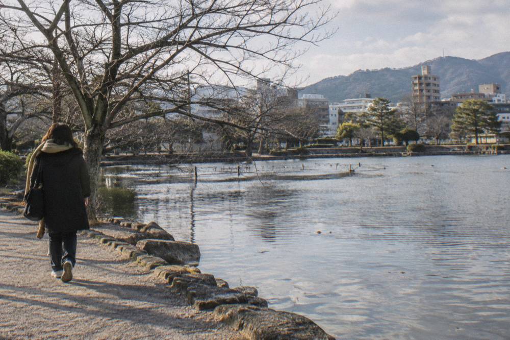 A visitor in a warm coat walking past bare winter trees beside a garden pond in Hiroshima