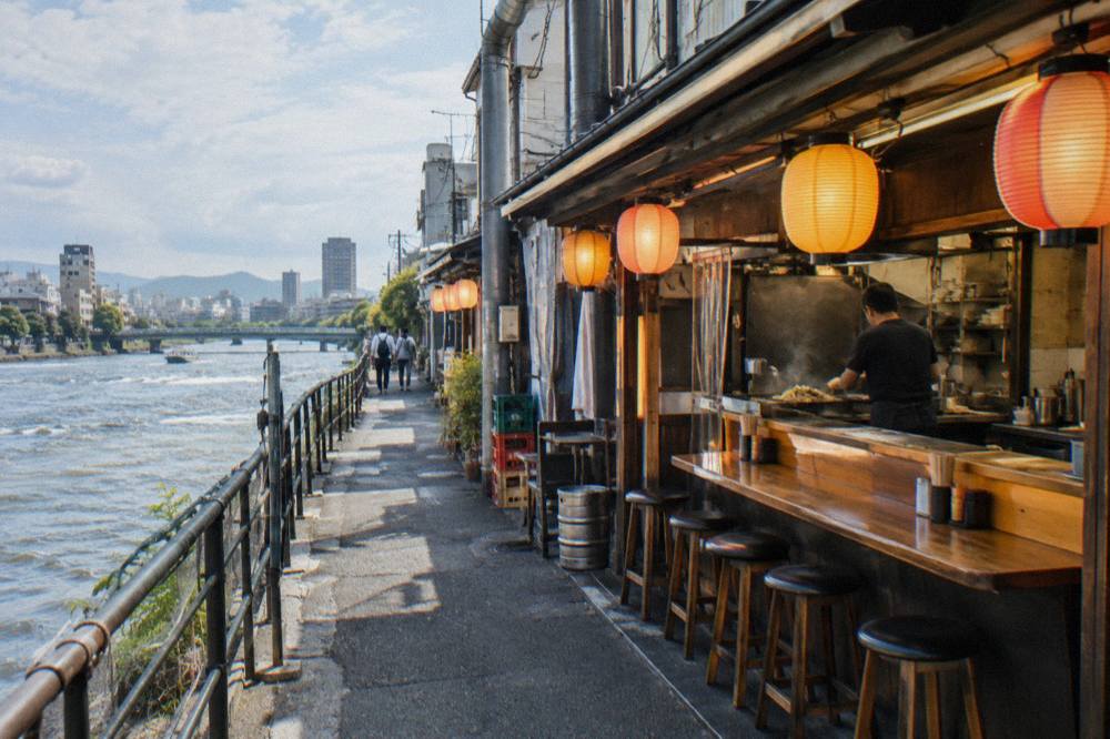 A small izakaya counter with lanterns glowing over stools and a cook working a flat grill