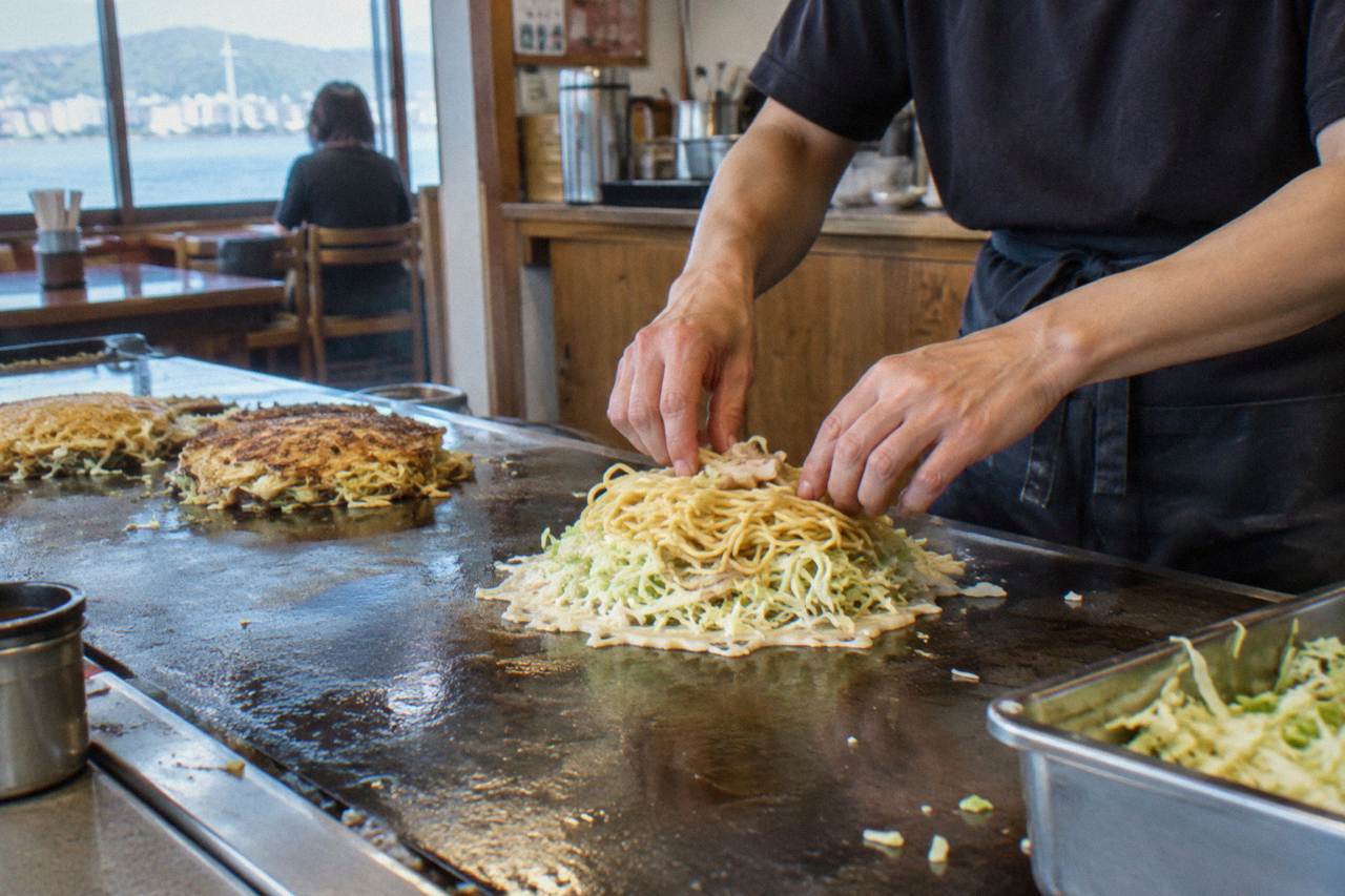 A chef layering cabbage and noodles on a thin batter base on a hot teppan griddle in a Hiroshima okonomiyaki restaurant