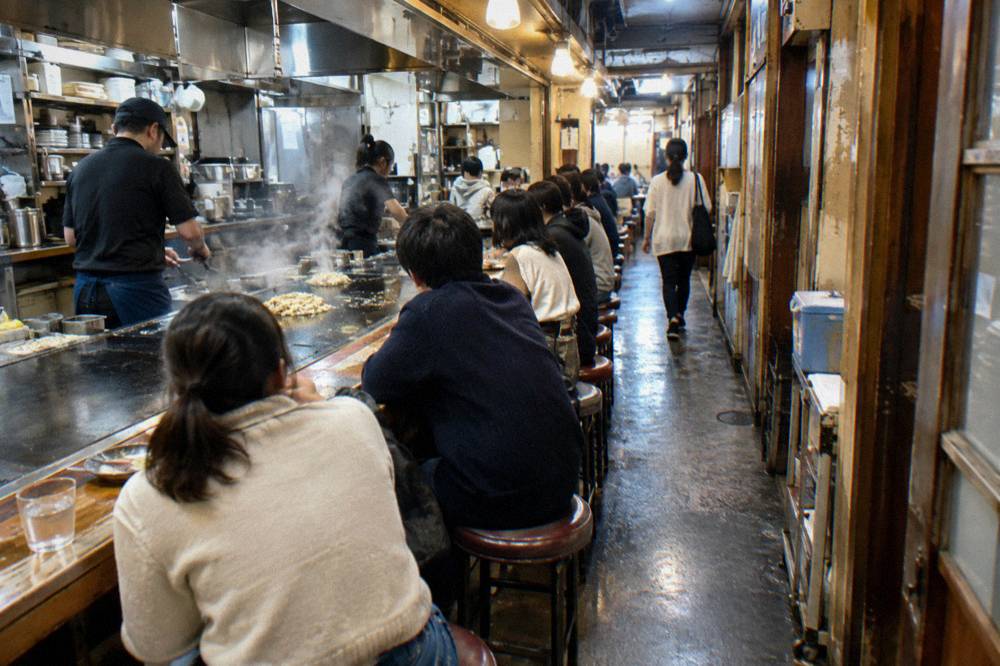 A narrow Okonomimura stall with customers seated at a counter around a wide flat griddle