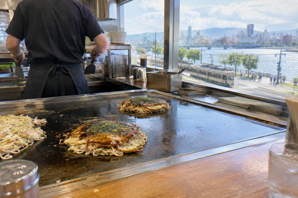 An okonomiyaki being cooked on a flat teppan griddle at a station restaurant counter