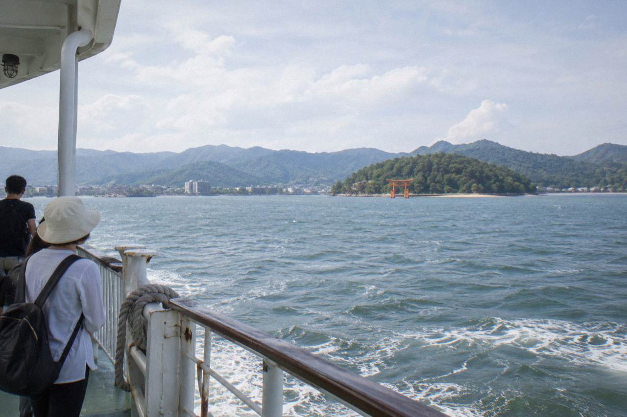 A red JR ferry crossing calm water toward the green island of Miyajima on a clear day