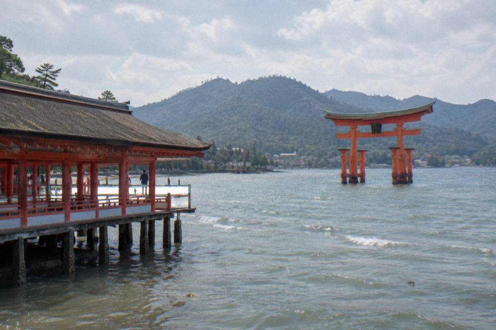 The view from a ferry deck of the red Great Torii gate rising from the sea beside forested hills
