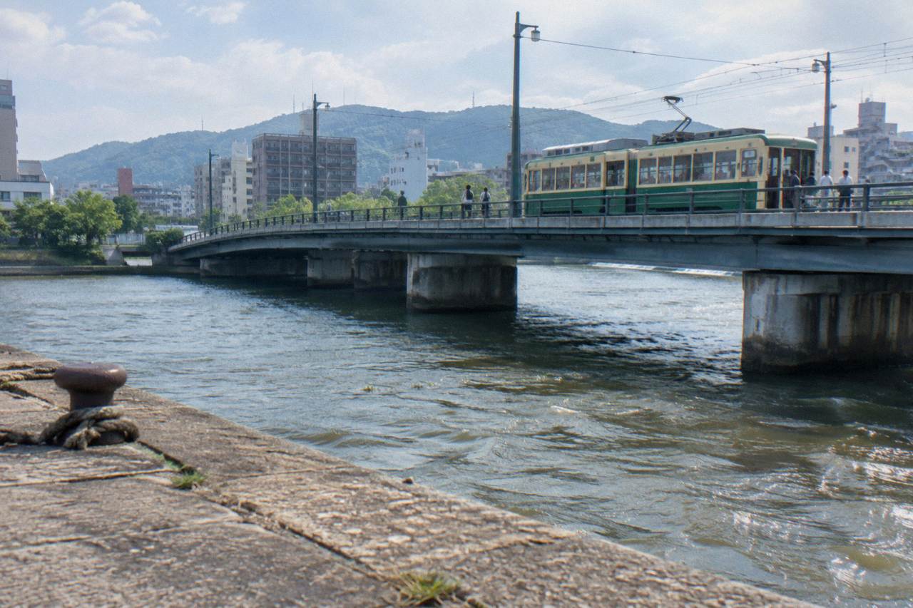 A green Hiroden streetcar crossing a bridge in central Hiroshima with passengers boarding