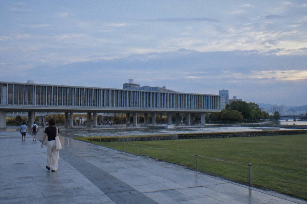 The pale concrete Hiroshima Peace Memorial Museum raised on pillars above a plaza at dusk, visitors walking below