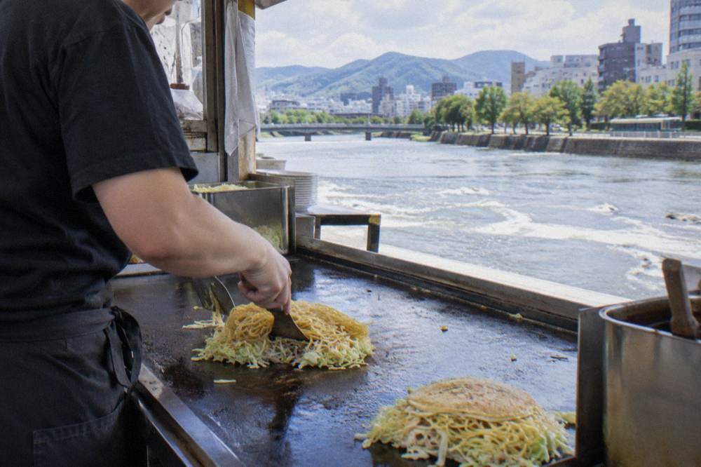 An okonomiyaki cook layering cabbage and noodles on a hot teppan in a Hiroshima stall