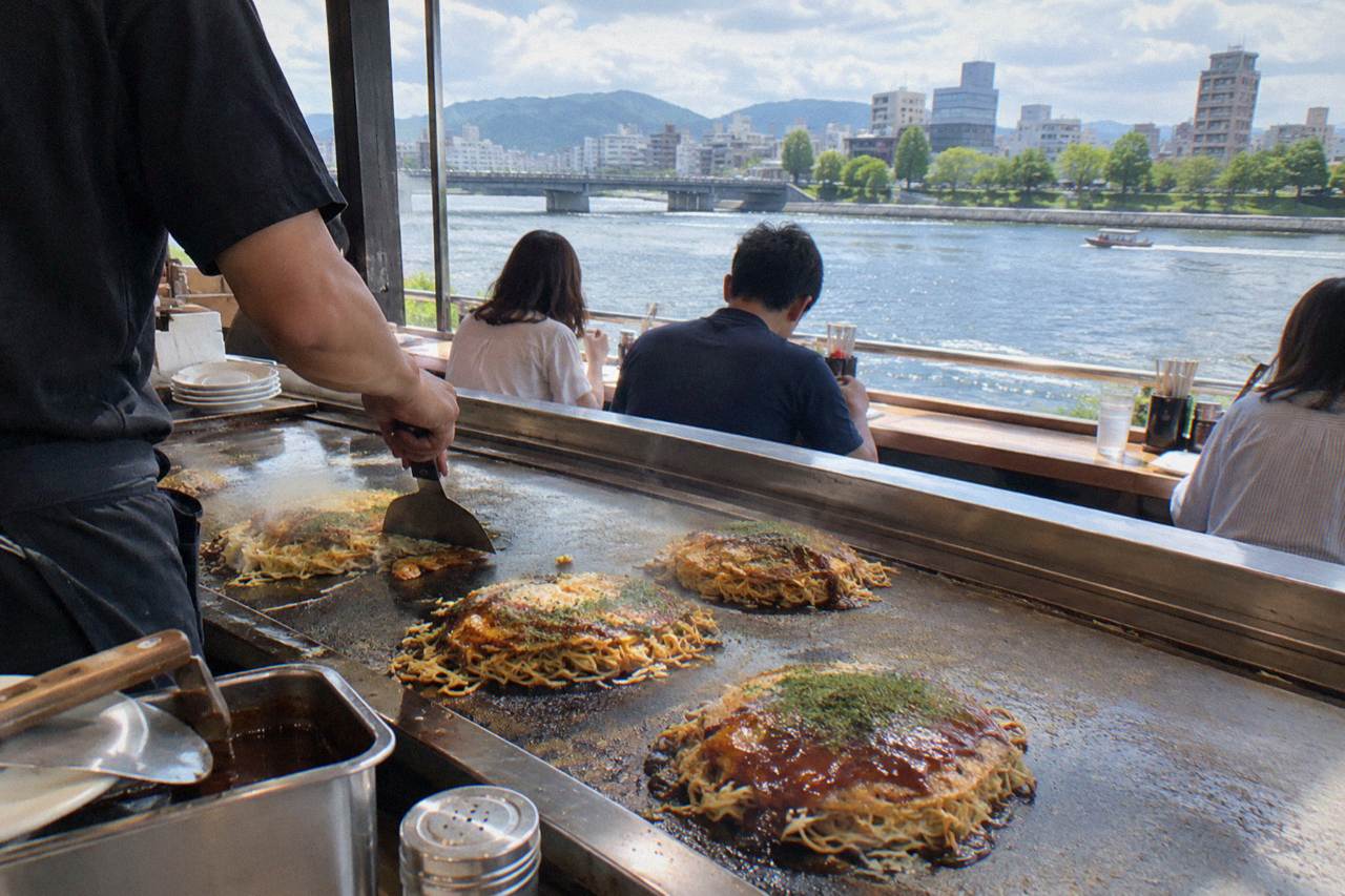 A hot iron griddle with Hiroshima-style okonomiyaki being cooked in front of a counter of diners