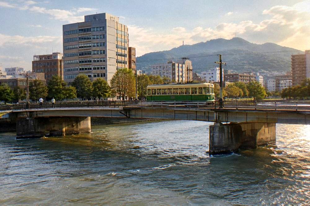 A Hiroden green streetcar crossing a bridge in central Hiroshima with office buildings behind it