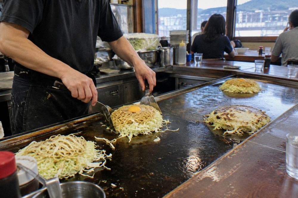 Chef layers cabbage, noodles and egg on a hot steel griddle while diners watch from counter seats