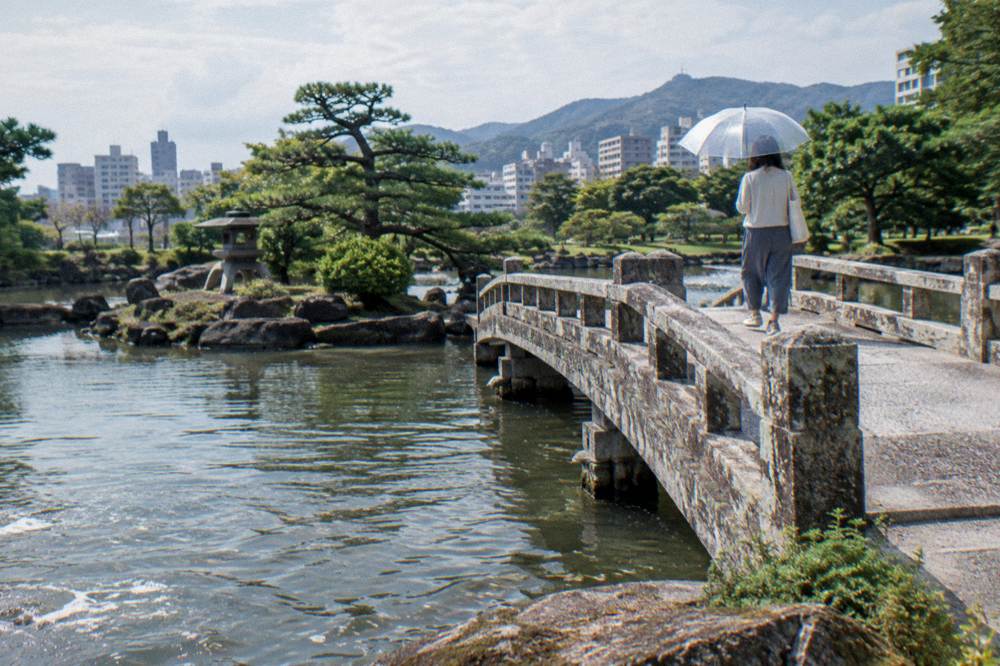A visitor with a clear umbrella crosses a stone bridge over a pond in a Japanese garden.