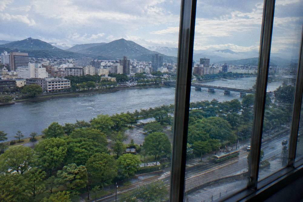 Rain streaks the tall windows of an observation deck overlooking a green park and river.