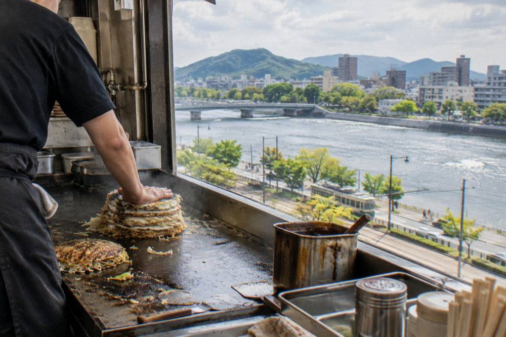 A cook presses a tall stack of okonomiyaki flat on the griddle while sauce waits nearby.