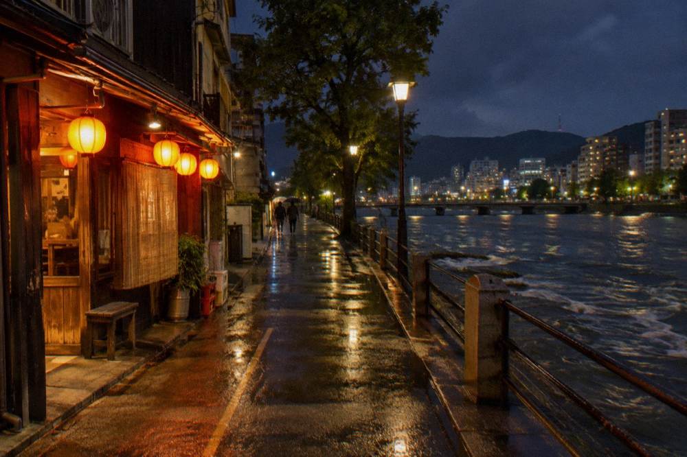 Warm lantern light spills from an izakaya doorway onto a wet downtown lane at night.