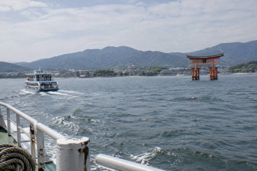 A ferry crosses open water toward a vermillion torii gate rising from the shallows.