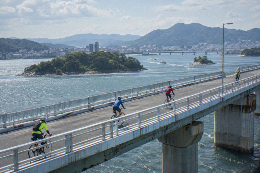 Cyclists in bright gear pedal a long bridge high above scattered islands and blue sea.