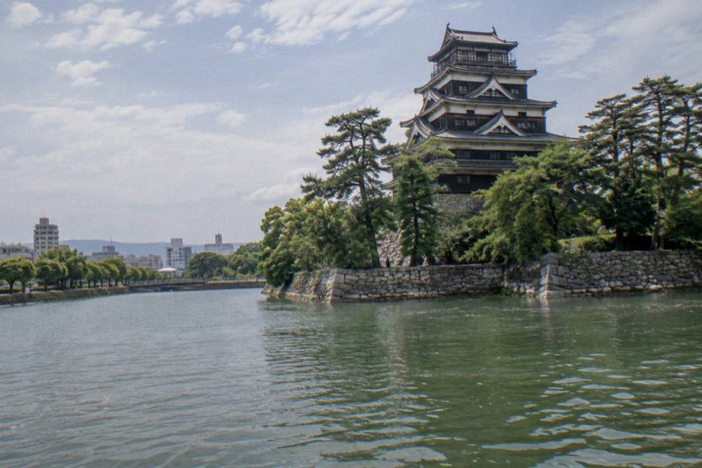 Hiroshima Castle keep seen across its moat, with stone walls and pine trees in view.
