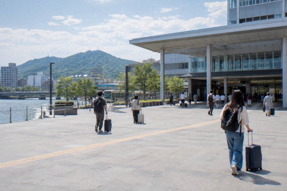 Travelers with rolling suitcases crossing the plaza in front of Hiroshima Station's modern south entrance.