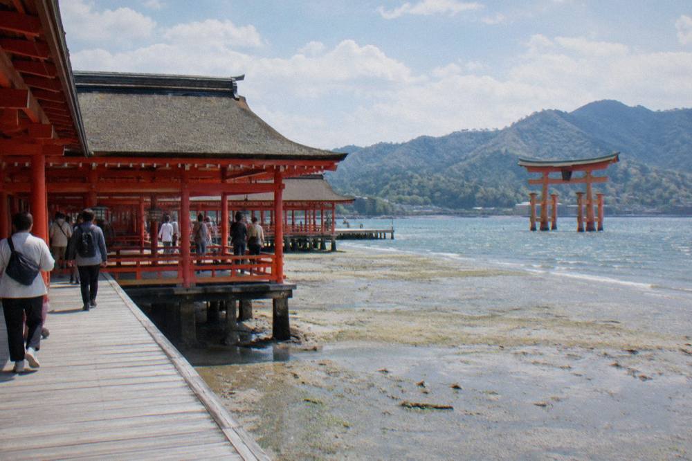 Visitors walking the wooden corridors of Itsukushima Shrine at low tide with the shoreline visible